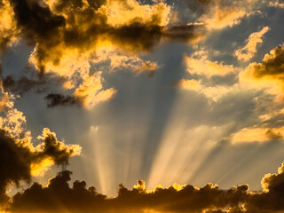 Dramatic sunset at end of summer, with sunbeams rising from behind shadowed clouds and illuminating others high above, as if to announce or preview something spectacular