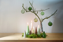 Alternative Advent Wreath Decoration With Four Different Candles, Small Artificial Christmas Trees And Green Glass Baubles On A Bare Branch Against A Light Wall, Copy Space