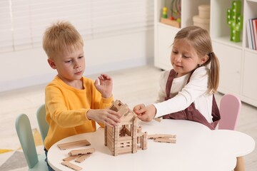 Sticker - Little boy and girl playing with wooden house at white table indoors. Children's toys