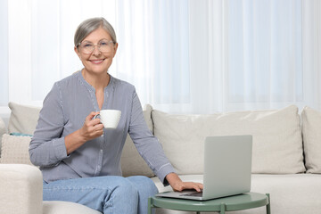 Poster - Beautiful senior woman with cup of drink using laptop at home, space for text