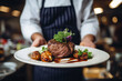 © MNStudio - Waiter holding a plate with grilled beef steak with roasted vegetables on a side. Serving fancy food in a restaurant.