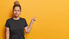Waist Up Shot Of Discontent Sulking Woman Frowns Face Indicates At Blank Copy Space With Frustrated Expression Dressed In Casual Black T Shirt Isolated On Yellow Background. Advertisement Concept