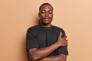 Calm dark skinned African American man embracing himself keeps eyes closed wears casual black t shirt enjoys wearing soft clothes loves himself stands against brown background. Self esteem concept