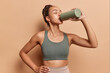 © WHstudio Leushin N - Sports and hydration concept. Sporty Latin with dark hair woman drinking fresh water from bottle sportswear top bra taking break after workout isolated over brown studio background feels thirsty