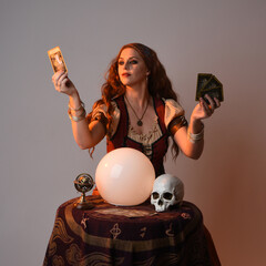 Wall Mural - close up portrait of beautiful red haired woman wearing a medieval fantasy fortune teller costume, looking into crystal ball reading the future at seance table. isolated on studio background 