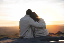 Back view of the happy couple in love sitting on top of a mountain enjoying a sunset landscape view