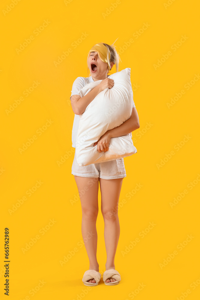 Young woman with pillow yawning on yellow background