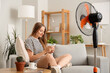 © Pixel-Shot - Young woman with blowing electric fan using mobile phone at home