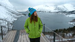 Lofoten islands, Senja region, Norway. Winter. Young girl in green jacket and blue hat stands on observation deck, looking away. In background snow-capped mountains, ocean Bay. Concept of tourism.