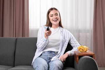 Poster - Happy woman switching TV channels at home. Nacho chips on wooden sofa armrest table