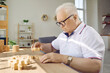 © Studio Romantic - Senior man playing puzzles to keep his brain young. Chubby old man in glasses sitting at desk at home and connecting wooden jigsaw puzzle pieces. Dementia and Alzheimer's disease prevention concept