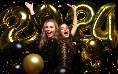 Happy gorgeous girls in stylish sexy party dresses holding gold 2024 balloons, having fun at New Year's Eve Party.