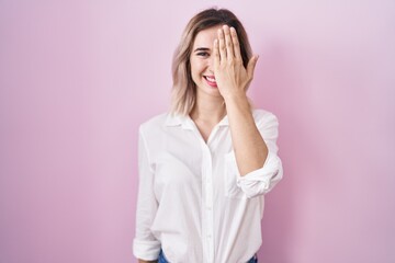 Poster - Young beautiful woman standing over pink background covering one eye with hand, confident smile on face and surprise emotion.