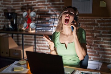 Wall Mural - Young beautiful woman working at the office at night speaking on the phone crazy and mad shouting and yelling with aggressive expression and arms raised. frustration concept.