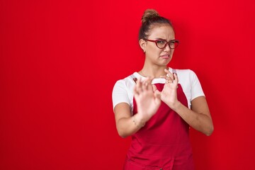 Canvas Print - Young hispanic woman wearing waitress apron over red background disgusted expression, displeased and fearful doing disgust face because aversion reaction. with hands raised