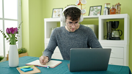 Wall Mural - Young hispanic man student using laptop taking notes wearing headphones at dinning room