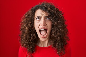 Sticker - Hispanic woman with curly hair standing over red background angry and mad screaming frustrated and furious, shouting with anger looking up.