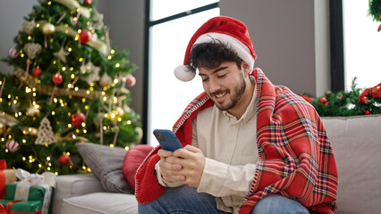 Wall Mural - Young hispanic man celebrating christmas using smartphone at home