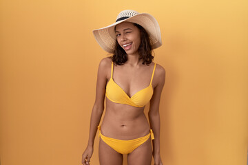 Poster - Young hispanic woman wearing bikini and summer hat winking looking at the camera with sexy expression, cheerful and happy face.