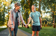 Cheerful modern senior couple outdoors in the park dressed in sportswear walking holding hands and laughing. Morning sun rays fall on smiling man's face..