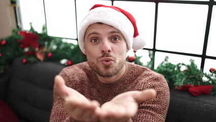 Poster - Young caucasian man sitting on sofa by christmas tree blowing a kiss at home