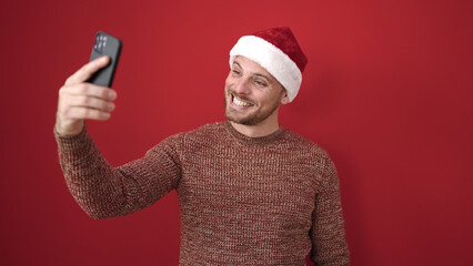 Sticker - Young caucasian man smiling taking selfie picture wearing christmas hat over isolated red background