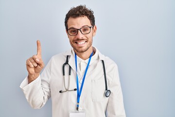 Wall Mural - Young hispanic man wearing doctor uniform and stethoscope showing and pointing up with finger number one while smiling confident and happy.