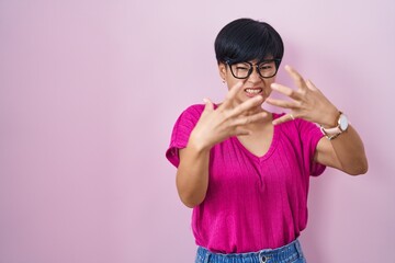 Poster - Young asian woman with short hair standing over pink background shouting frustrated with rage, hands trying to strangle, yelling mad