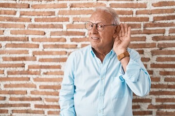 Canvas Print - Senior man with grey hair standing over bricks wall smiling with hand over ear listening an hearing to rumor or gossip. deafness concept.