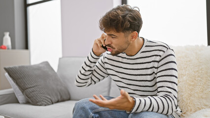 Sticker - Stressed-out young arab man furiously arguing over phone, sitting on his living room sofa, evoking an intense domestic tension