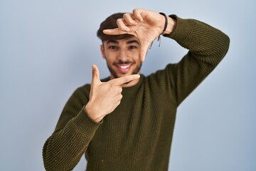 Sticker - Arab man with beard standing over blue background smiling making frame with hands and fingers with happy face. creativity and photography concept.