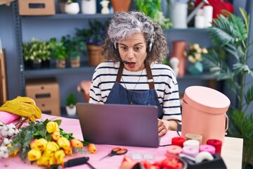 Wall Mural - Middle age woman with grey hair working at florist with laptop scared and amazed with open mouth for surprise, disbelief face