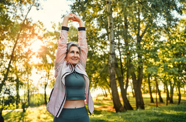 Wall Mural - Active aging and outdoor workout. Enduring mature woman with grey hair doing regular workout on fresh air. Positive lady wearing sport outfit performing arms exercises and strengthening back.