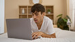 © Krakenimages.com - Handsome young hispanic man lounging comfortably in a cozy bed, engrossed in his laptop, enjoying a relaxing morning in his bedroom at home.