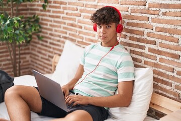 Poster - Young hispanic teenager using laptop and headphones sitting on bed at bedroom