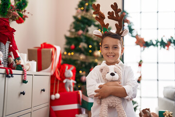 Sticker - Adorable hispanic toddler hugging teddy bear standing by christmas tree at home