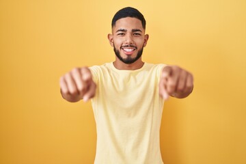 Young hispanic man standing over yellow background pointing to you and the camera with fingers, smiling positive and cheerful