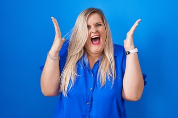 Poster - Caucasian plus size woman standing over blue background celebrating crazy and amazed for success with arms raised and open eyes screaming excited. winner concept