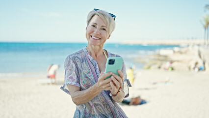 Wall Mural - Middle age blonde woman tourist smiling confident using smartphone at the beach