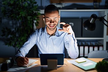 Canvas Print - Young hispanic man working at the office at night smiling and confident gesturing with hand doing small size sign with fingers looking and the camera. measure concept.