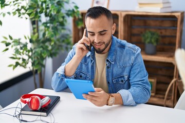 Poster - Young hispanic man using touchpad and talking on smartphone at home