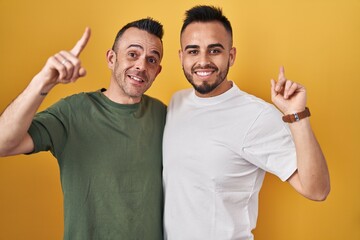 Sticker - Homosexual couple standing over yellow background smiling amazed and surprised and pointing up with fingers and raised arms.