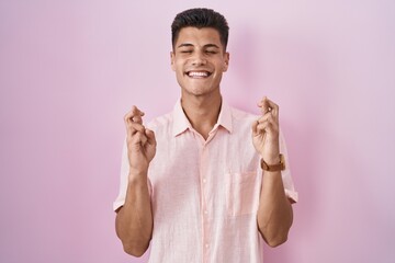 Canvas Print - Young hispanic man standing over pink background gesturing finger crossed smiling with hope and eyes closed. luck and superstitious concept.