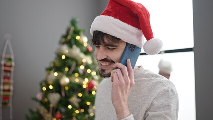 Canvas Print - Young hispanic man speaking on the phone by christmas tree at home