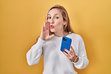 Wall Mural - Young blonde woman using smartphone typing message hand on mouth telling secret rumor, whispering malicious talk conversation