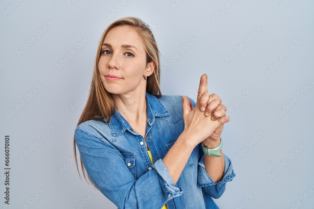 Young blonde woman standing over blue background holding symbolic gun with hand gesture, playing killing shooting weapons, angry face