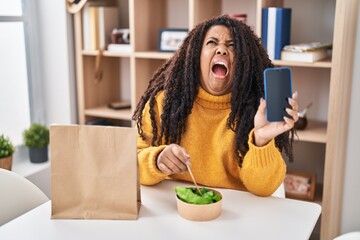 Wall Mural - Plus size hispanic woman eating take away salad holding smartphone angry and mad screaming frustrated and furious, shouting with anger looking up.