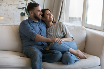 Wall Mural - Happy dating couple enjoying relaxation, leisure time at cozy home, discussing romantic plans, dreaming together, looking at window away, holding hands, hugging with love, affection