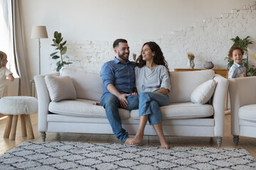 Wall Mural - Couple of happy parents enjoying being at new cozy stylish home, sitting on couch, laughing. Two active little sibling kids running around cheerful mom and dad, resting on sofa in modern interior