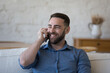 © fizkes - Happy cheerful millennial mobile phone user man enjoying call, conversation, feeling joy, talking on smartphone, sitting on home couch, looking away, enjoying leisure, communication,